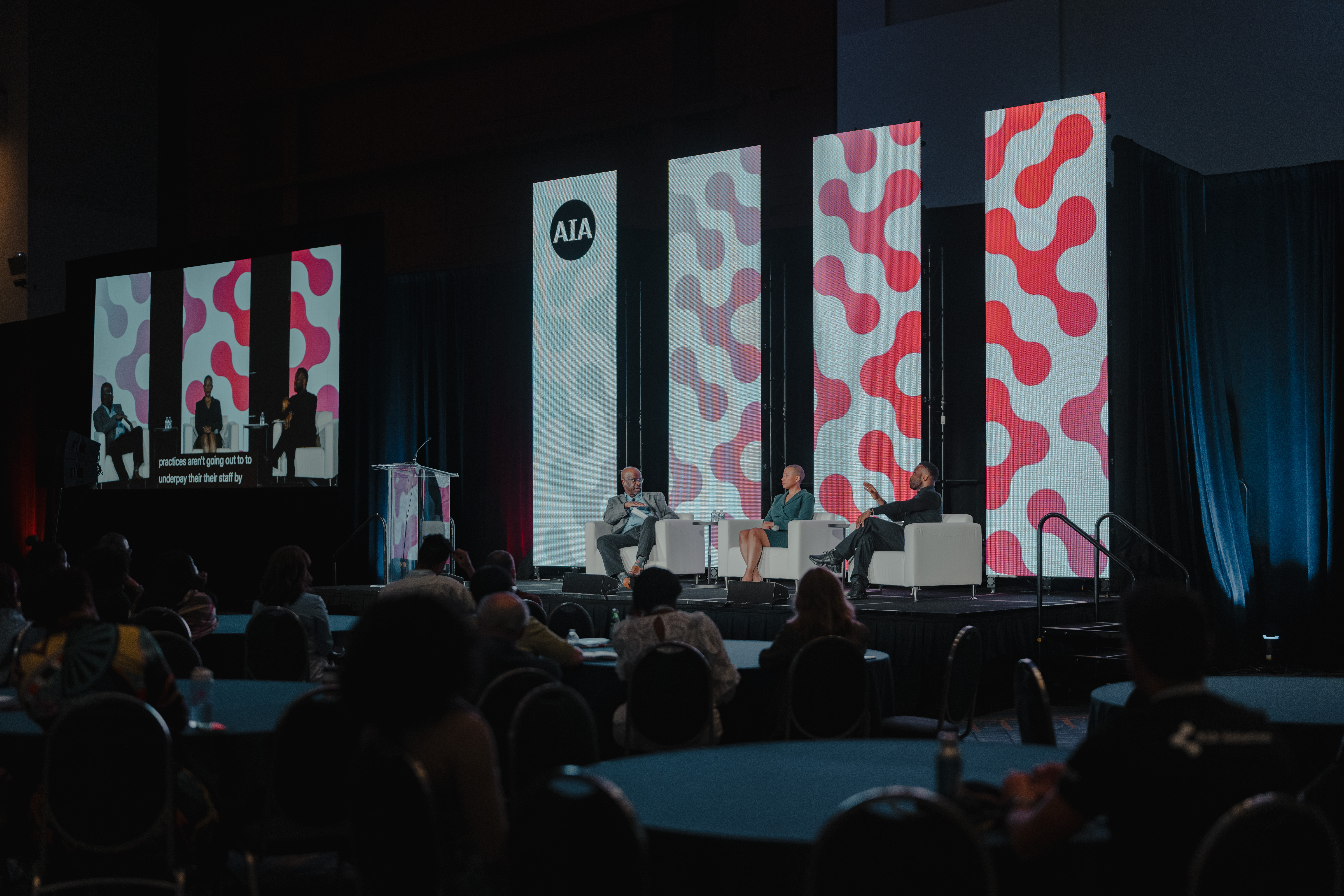 A panel discussion at an AIA (American Institute of Architects) event, featuring three panelists seated on stage in front of large screens with abstract red and gray designs. The event is taking place in a large conference room, with attendees seated at round tables. A video screen to the left displays the panelists, along with captions summarizing their discussion.