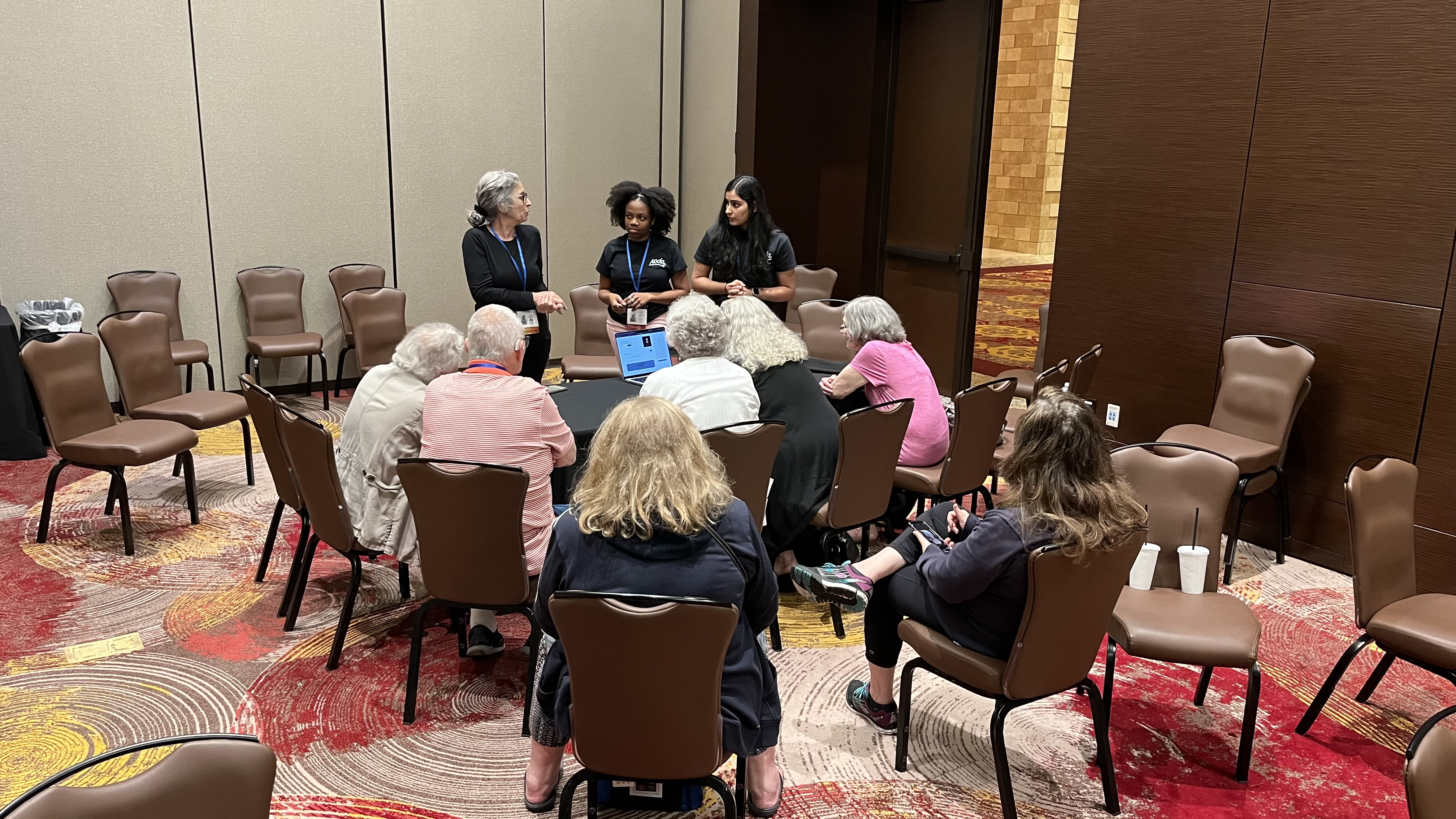 A group of older adults seated in a semi-circle in a conference room, listening to a presentation by three women standing at the front. One of the presenters is speaking while the other two stand by, and a laptop is open on the table displaying information. The room has patterned red and yellow carpeting and brown chairs arranged around the room.