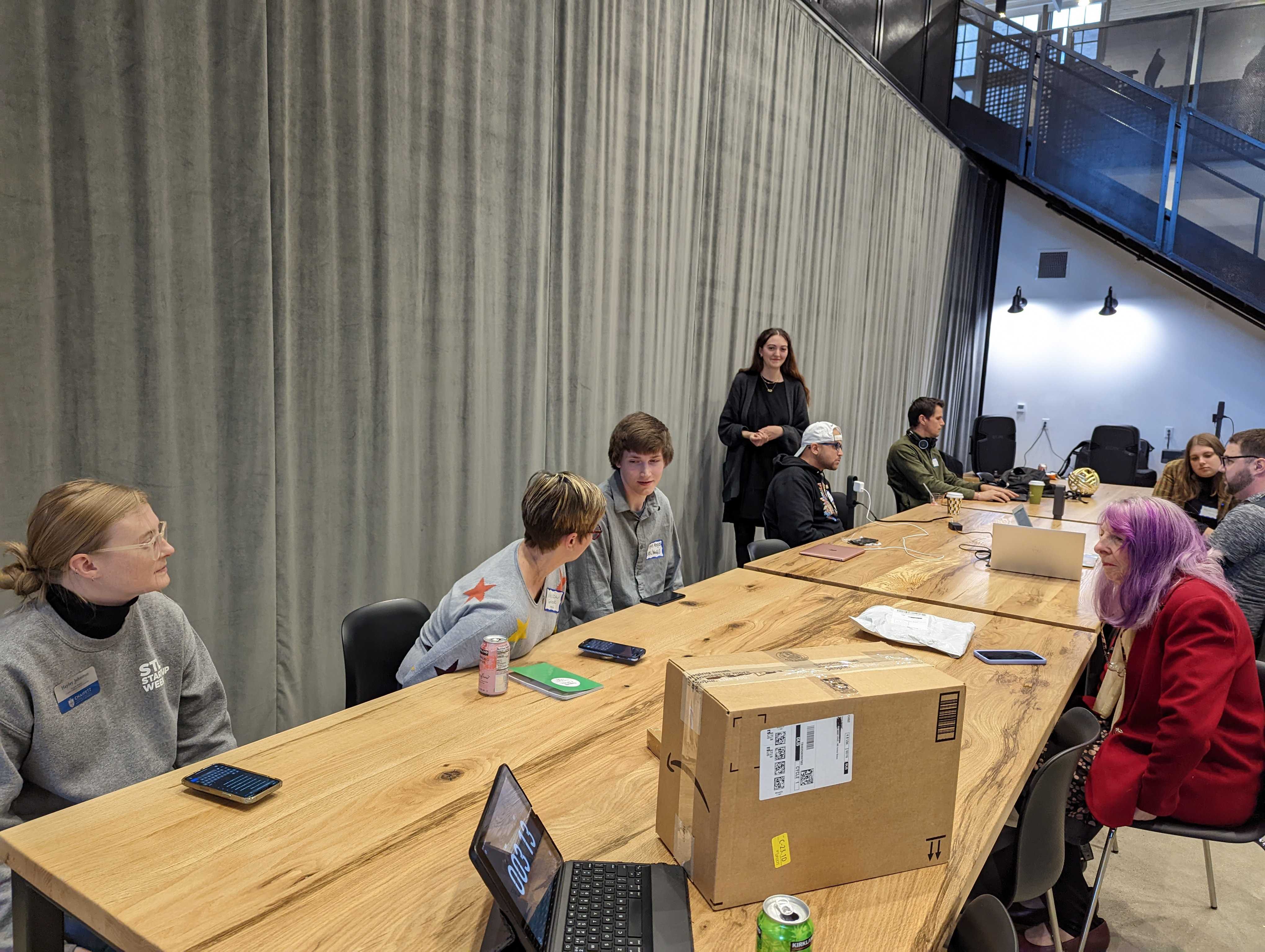 A group of people seated around a large wooden table during a workshop or meeting. Some are using laptops, while others have drinks and notebooks in front of them. A woman stands at the end of the table, appearing to lead the session. One participant has vibrant purple hair, and the room features modern decor with a high ceiling and gray curtains along one wall.